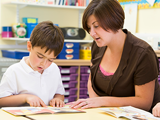 Teacher and student sitting a desk while reading a book together.