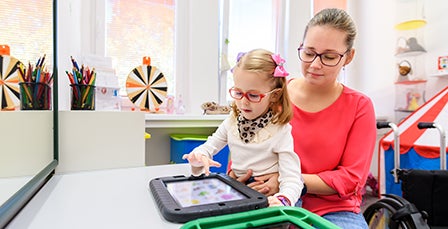 Woman holding child while sitting at a desk working on a tablet.