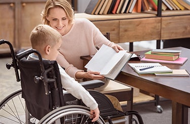 Boy in wheelchair working with teacher who is holding a book for him.