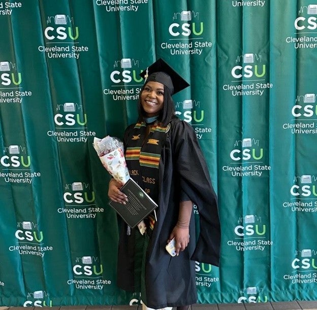 Student wearing graduation robe, holding flowers standing in front of a CSU banner